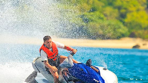 Fahrer mit oranger Schwimmweste auf einem Jetski, im Hintergrund Strand und Palmen bei Koh Samui